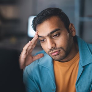 A stressed cybersecurity professional looking at a laptop screen with complex code and alerts.